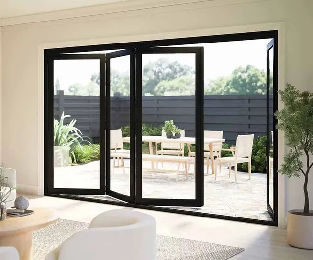 Dark grey aluminium bi-fold doors folded open from a living room onto a paved terrace with garden furniture